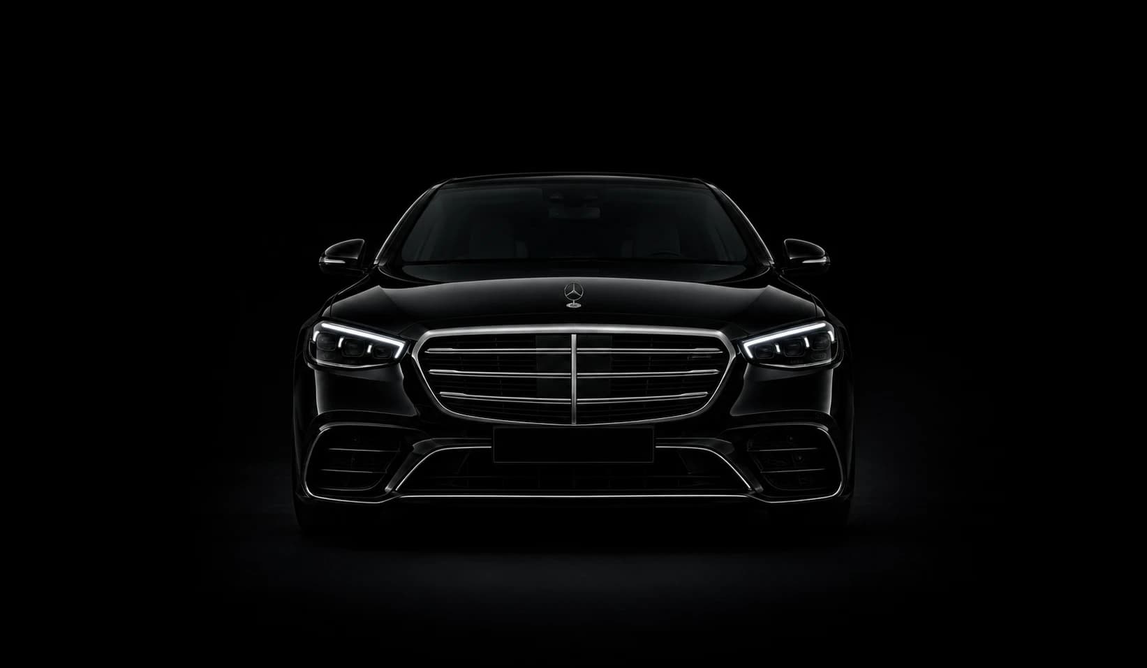 Black Mercedes-Benz S-Class saloon, front view, headlights on, against a black studio background