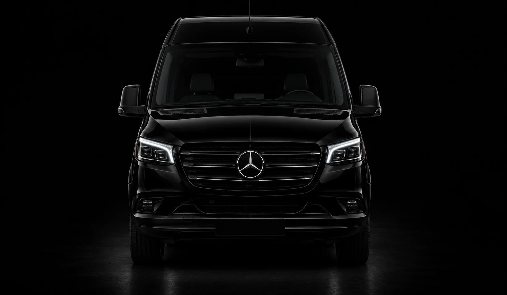 Black Mercedes-Benz Sprinter minibus, front view, headlights on, against a black studio background